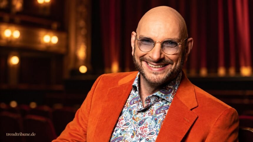 Ralph Morgenstern mit orangefarbenem Sakko vor einer gestalteten Theaterkulisse